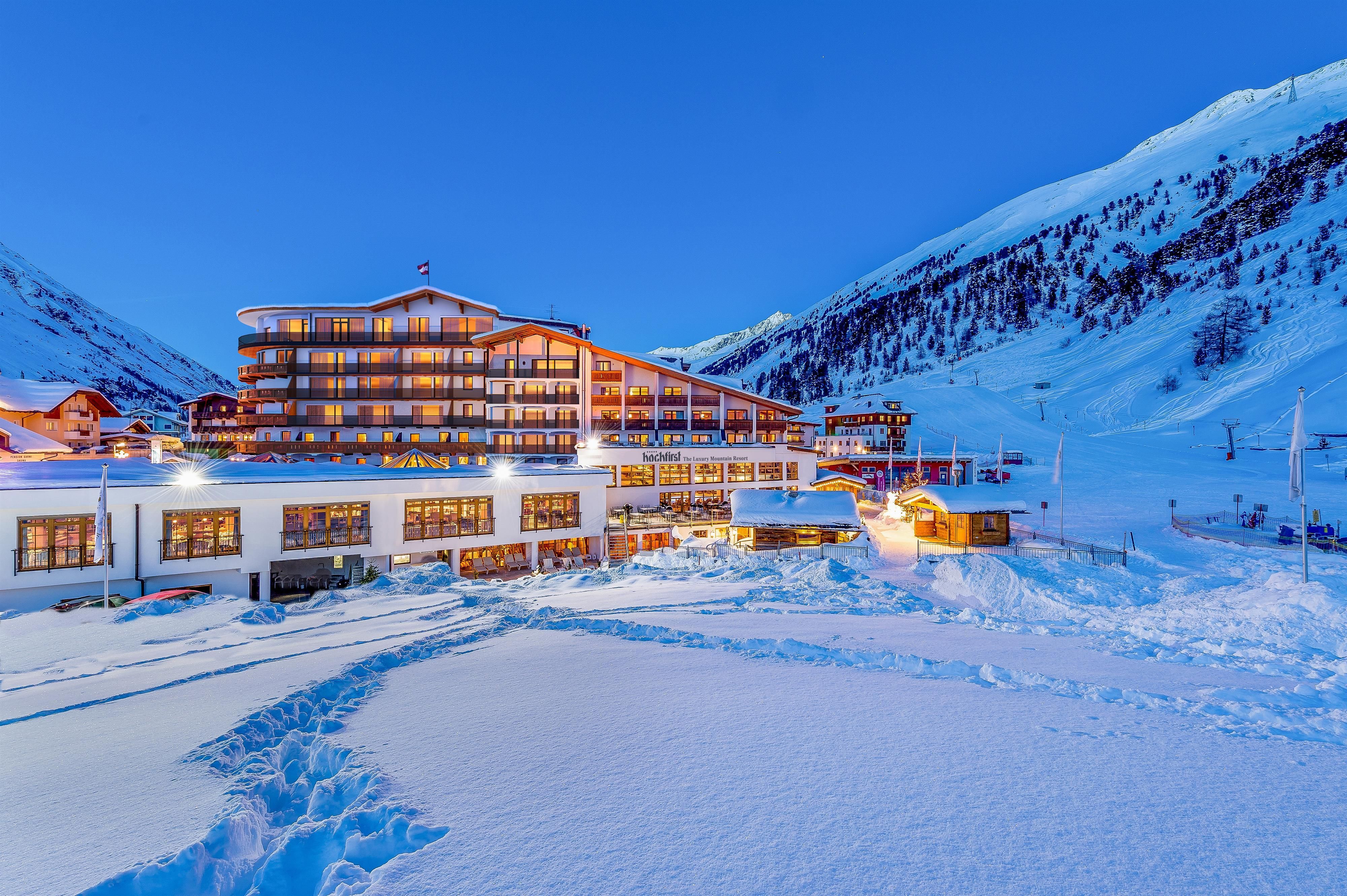 A modern hotel in the midst of a snow-covered landscape. The surroundings are surrounded by high mountains and the sky is clear and blue.