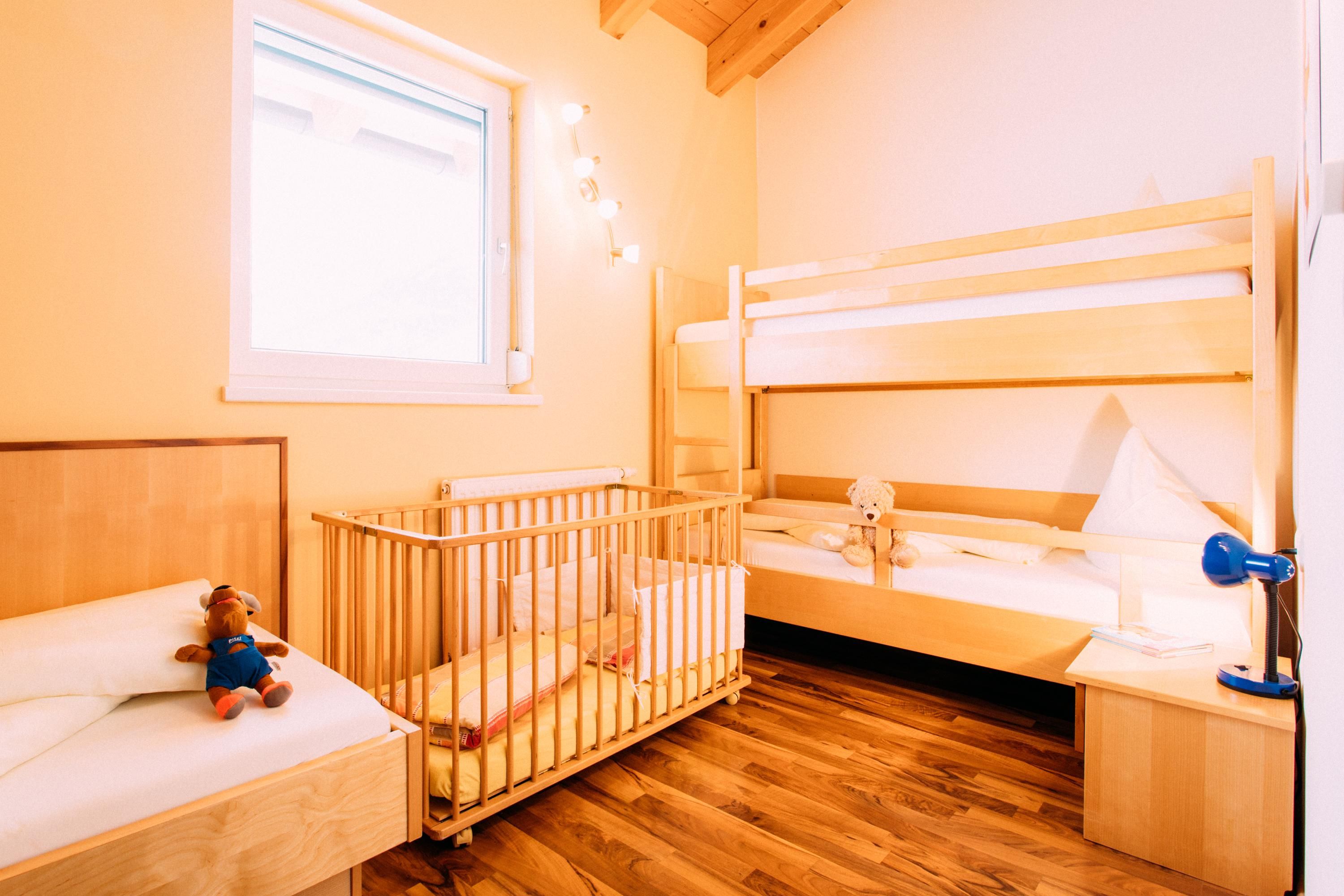 A bright children's room with a loft bed, a crib, and a single bed. There is wooden flooring and a large window opening with natural light.