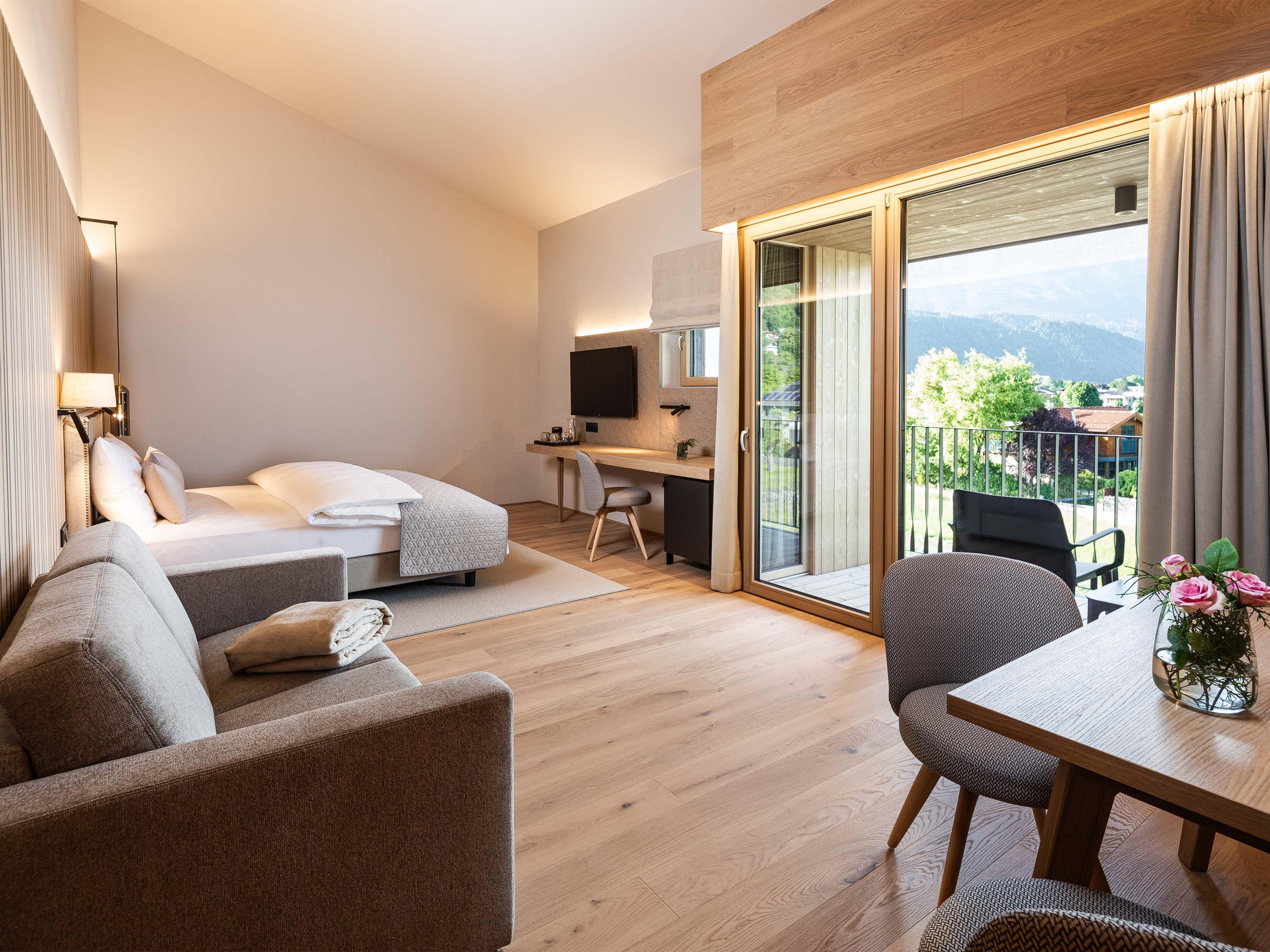 A modern room with light wooden furniture and large windows. There is a balcony with a view of the landscape and a cozy seating area.