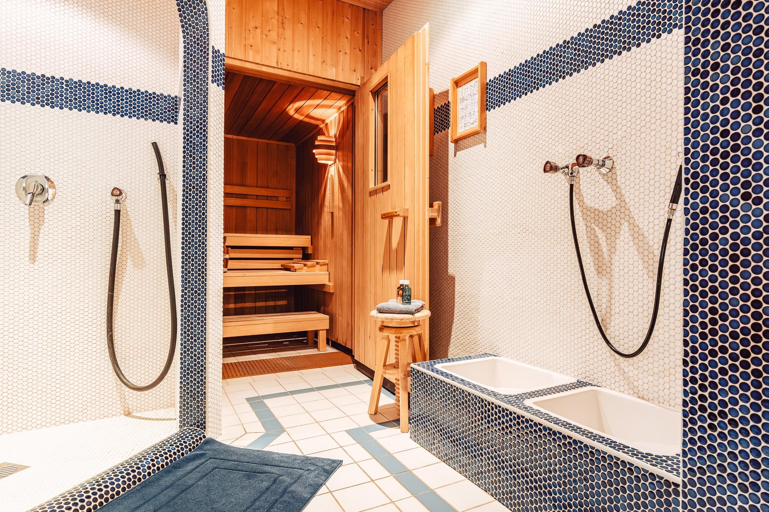 A modern bathroom with a sauna and beautiful blue tiles. Two shower areas and a wooden chair complete the relaxing ambiance.