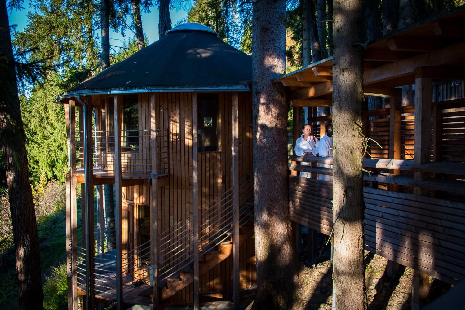 A modern wooden house stands among trees in nature. Two people are on the porch enjoying the view.