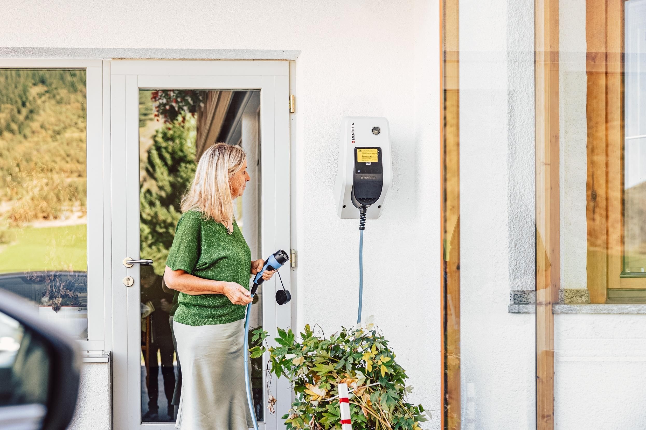 A woman is standing next to a wall charging station for electric cars. She is holding a charging cable in her hand and is looking at the door of the house.