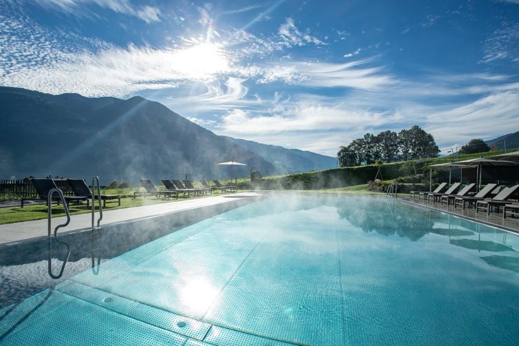 A beautiful pool with reflections of the sun and surrounding loungers. In the background, gentle hills and a clear blue sky can be seen.