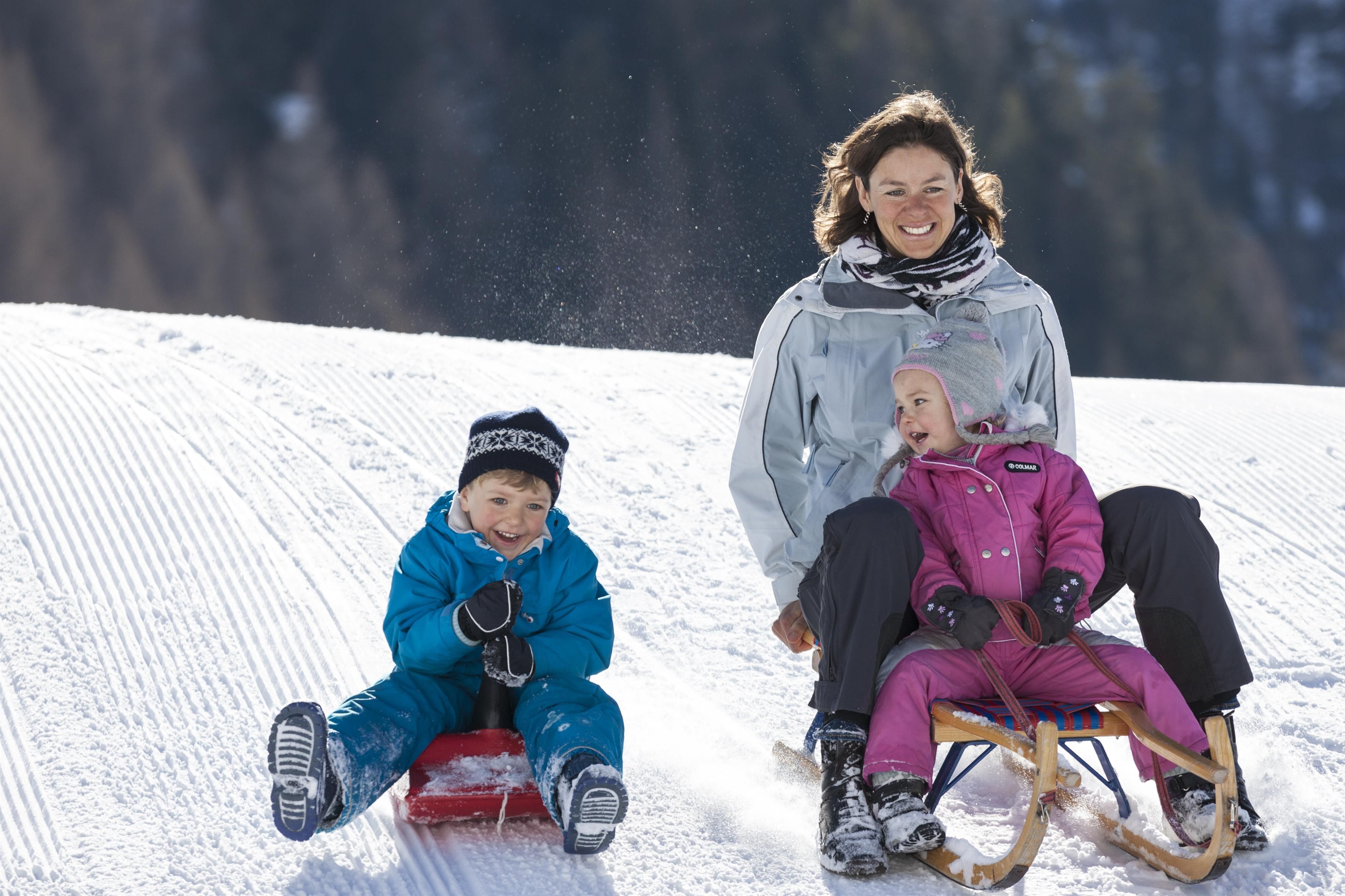A mother is sliding down a snowy slope with two children on a sled. The sun is shining and the surroundings are beautifully wintry.
