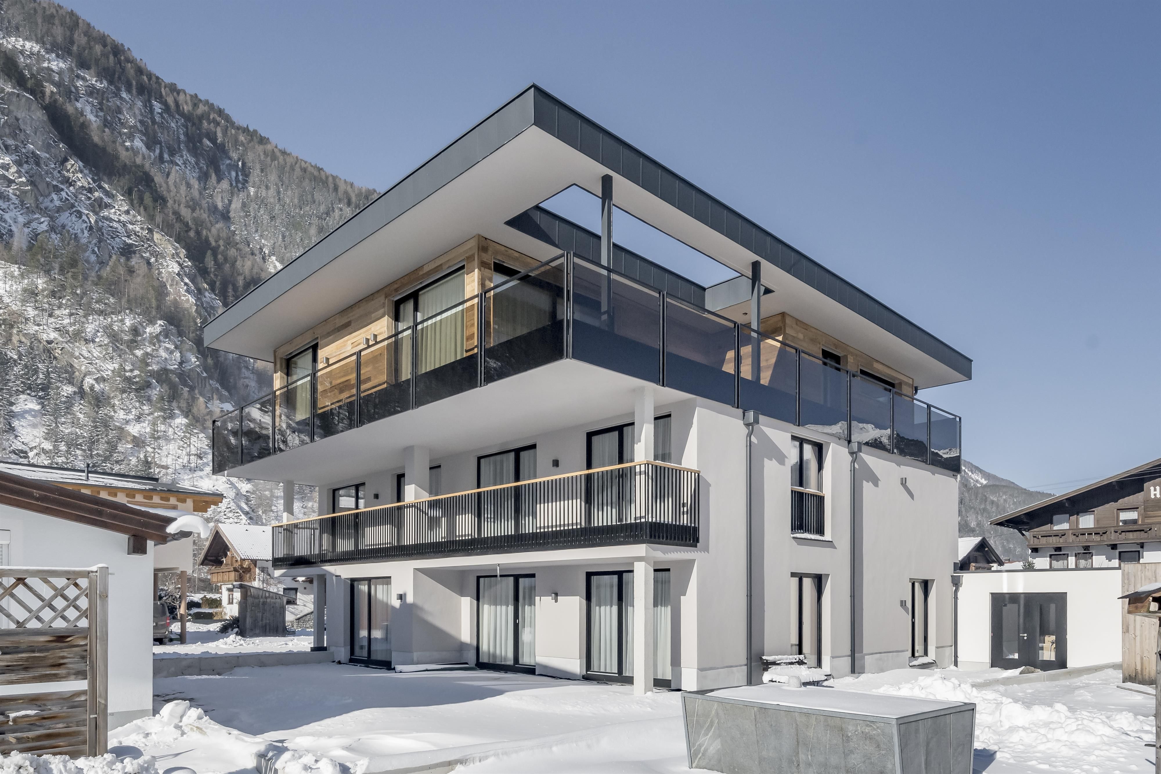 A modern building with several balconies in a snow-covered landscape. In the background, mountains are visible and the sky is clear.