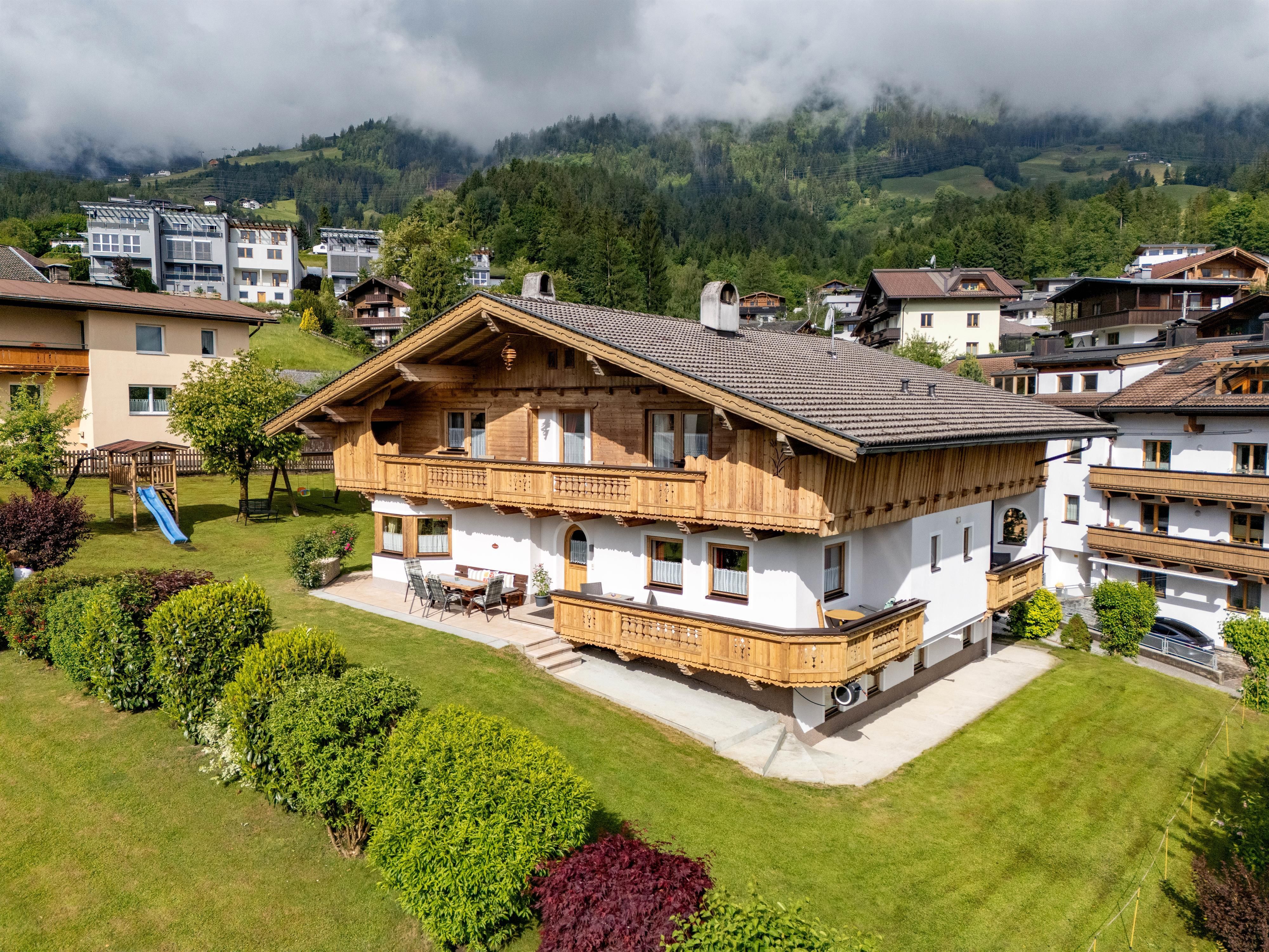 A charming alpine house with wooden cladding in a green setting. In the background, mountains and other buildings are visible.