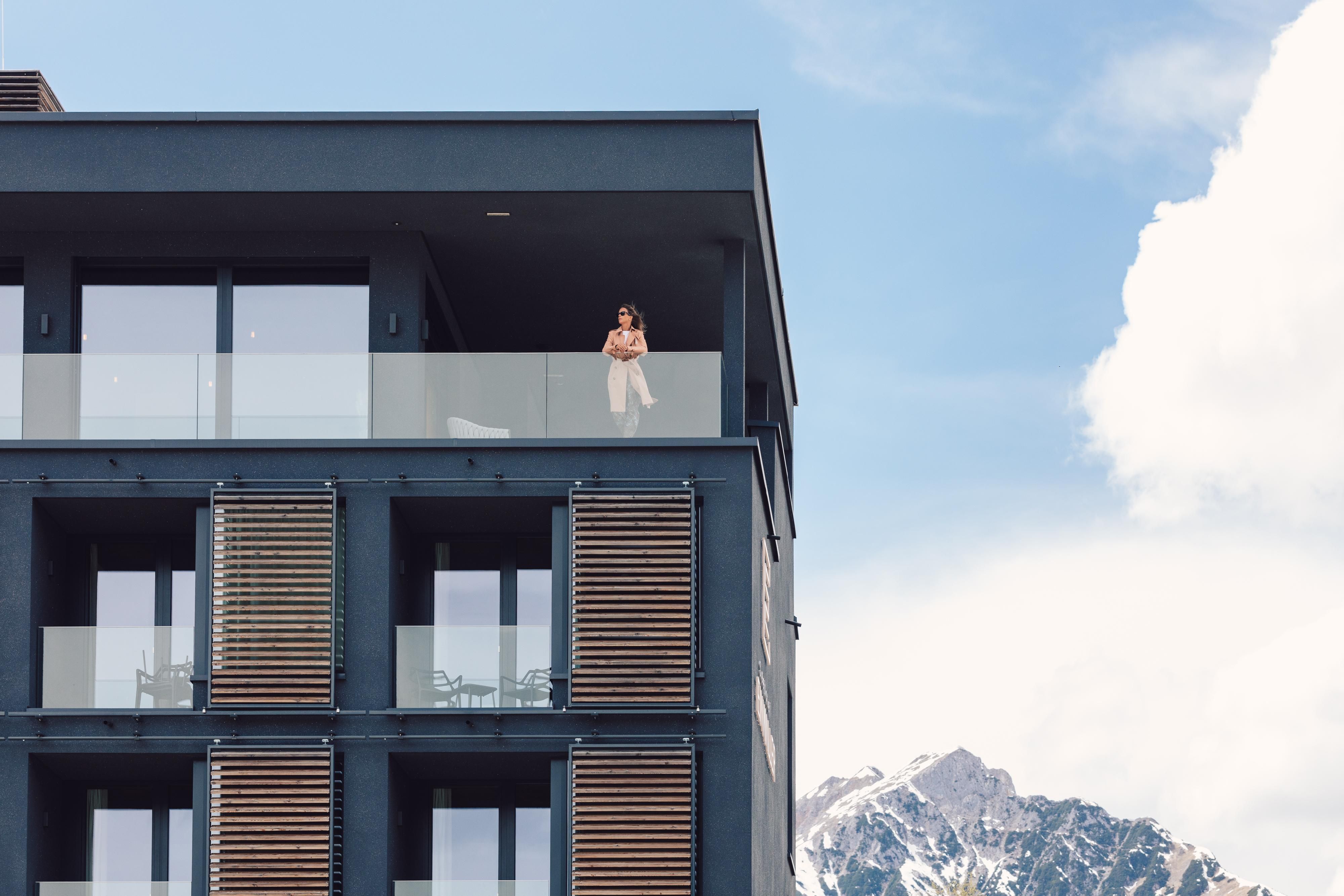 A modern building with balconies and large windows. In the background, impressive mountains and a blue sky can be seen.