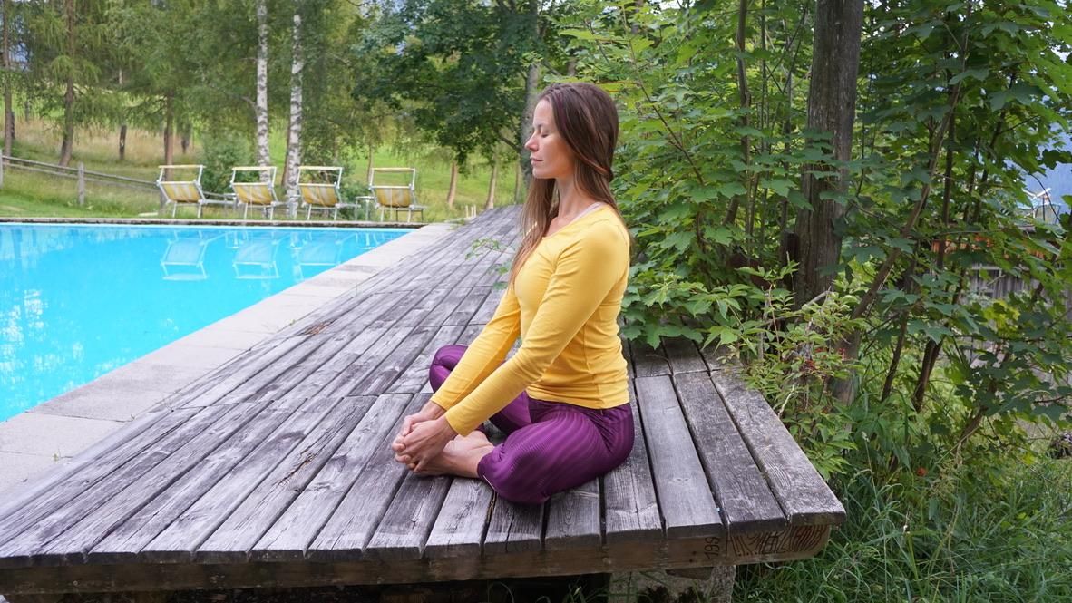 A meditating woman sits on a wooden platform next to a pool. She wears a yellow top and purple pants, surrounded by a serene garden.