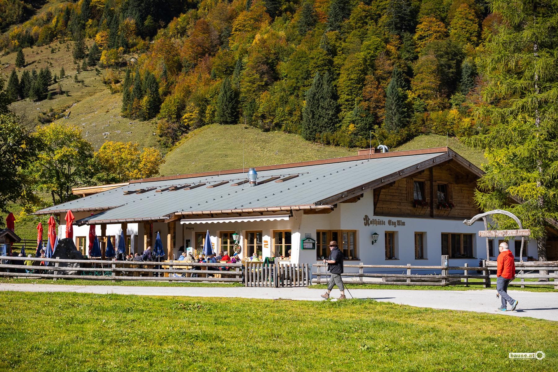 A cozy restaurant situated in a picturesque landscape. The surroundings showcase colorful autumn trees and walkers on a beautiful path.