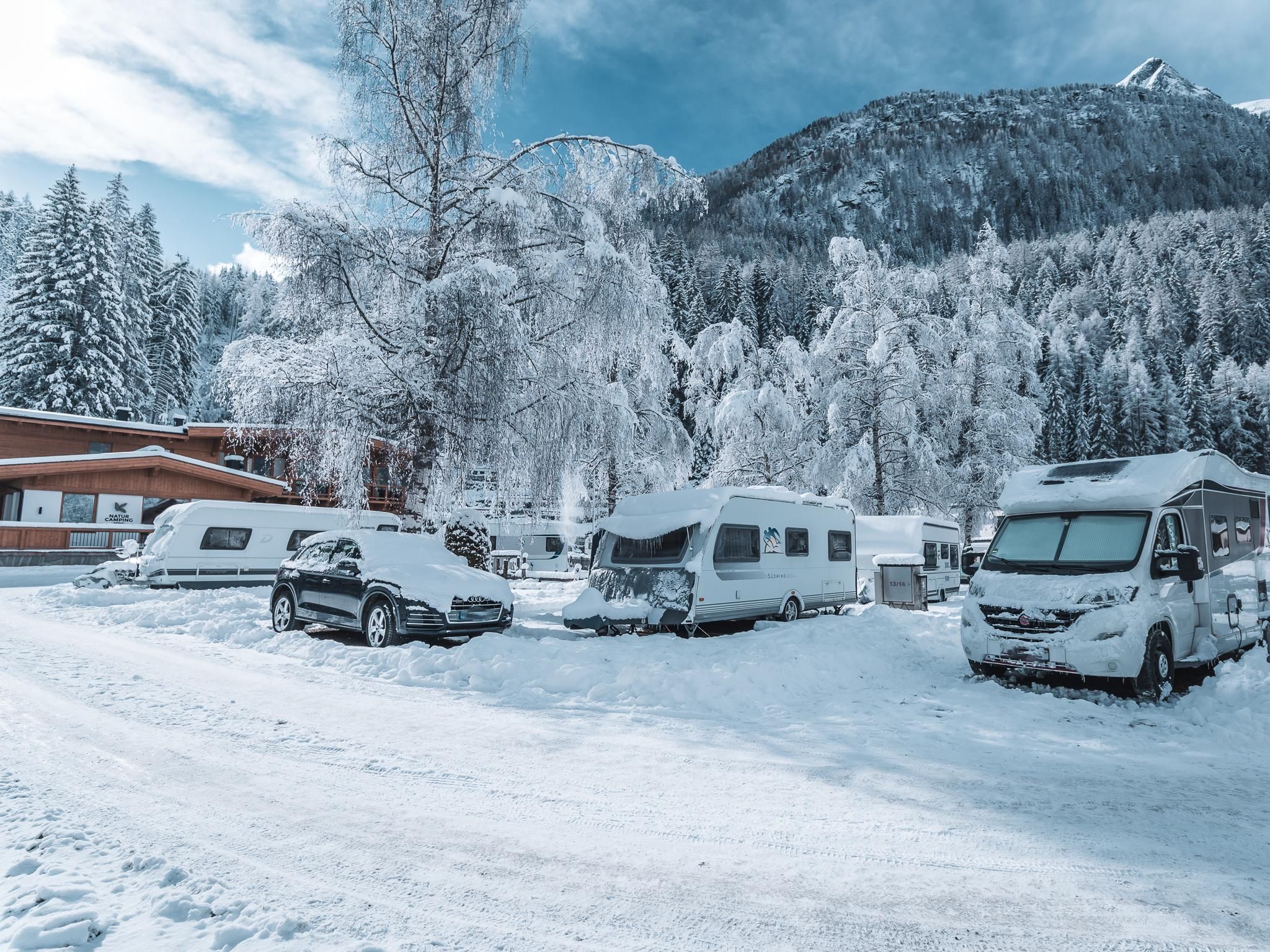 A winter campsite with several RVs covered in snow. In the background, snow-covered mountains and trees can be seen.