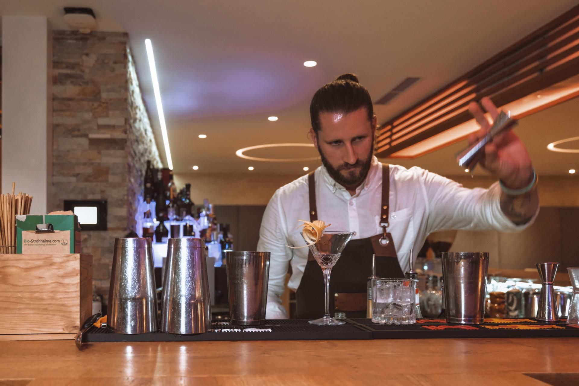 A bartender mixes cocktails at a modern bar. In the foreground are several shakers and glasses.