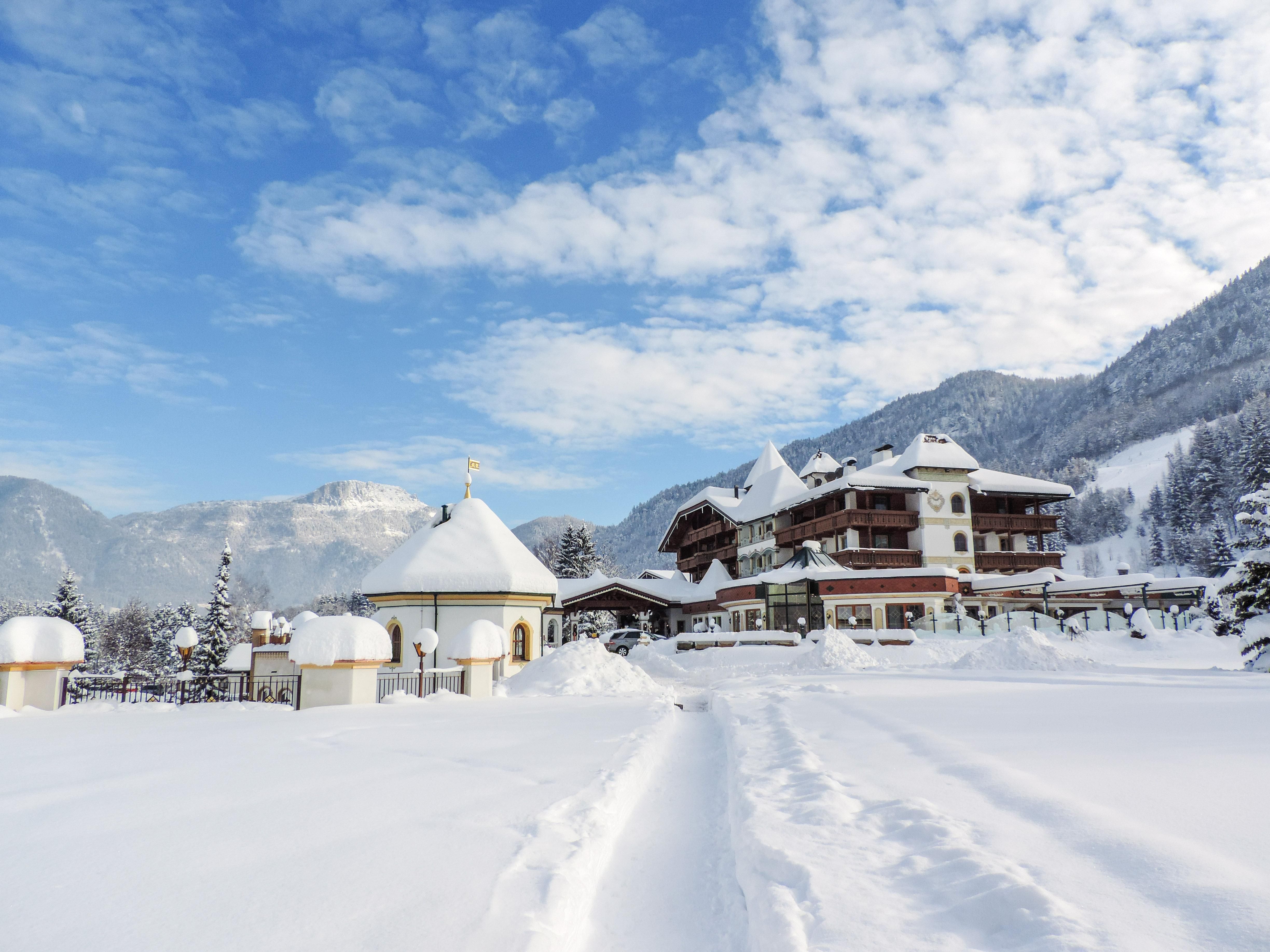 A picturesque winter landscape with plenty of snow and a classic building in the background. The sky is blue with some clouds, and the mountains are visible in the distance.