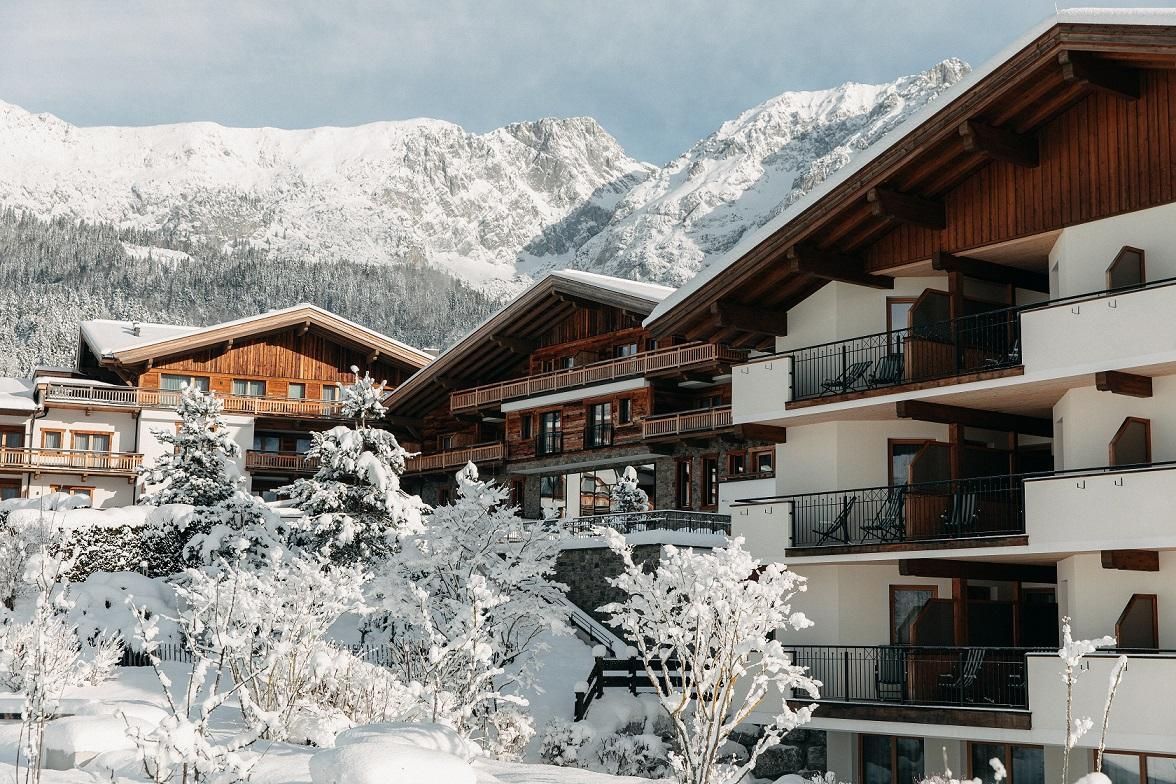 A picturesque winter landscape with snow-covered cabins and tall mountains in the background. The houses have wood-clad facades and are surrounded by snow-covered trees.
