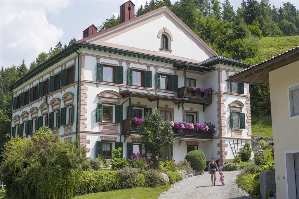 A large, elegant building with colorful flower balconies and green shutters. A person walks on a paved path towards the house.