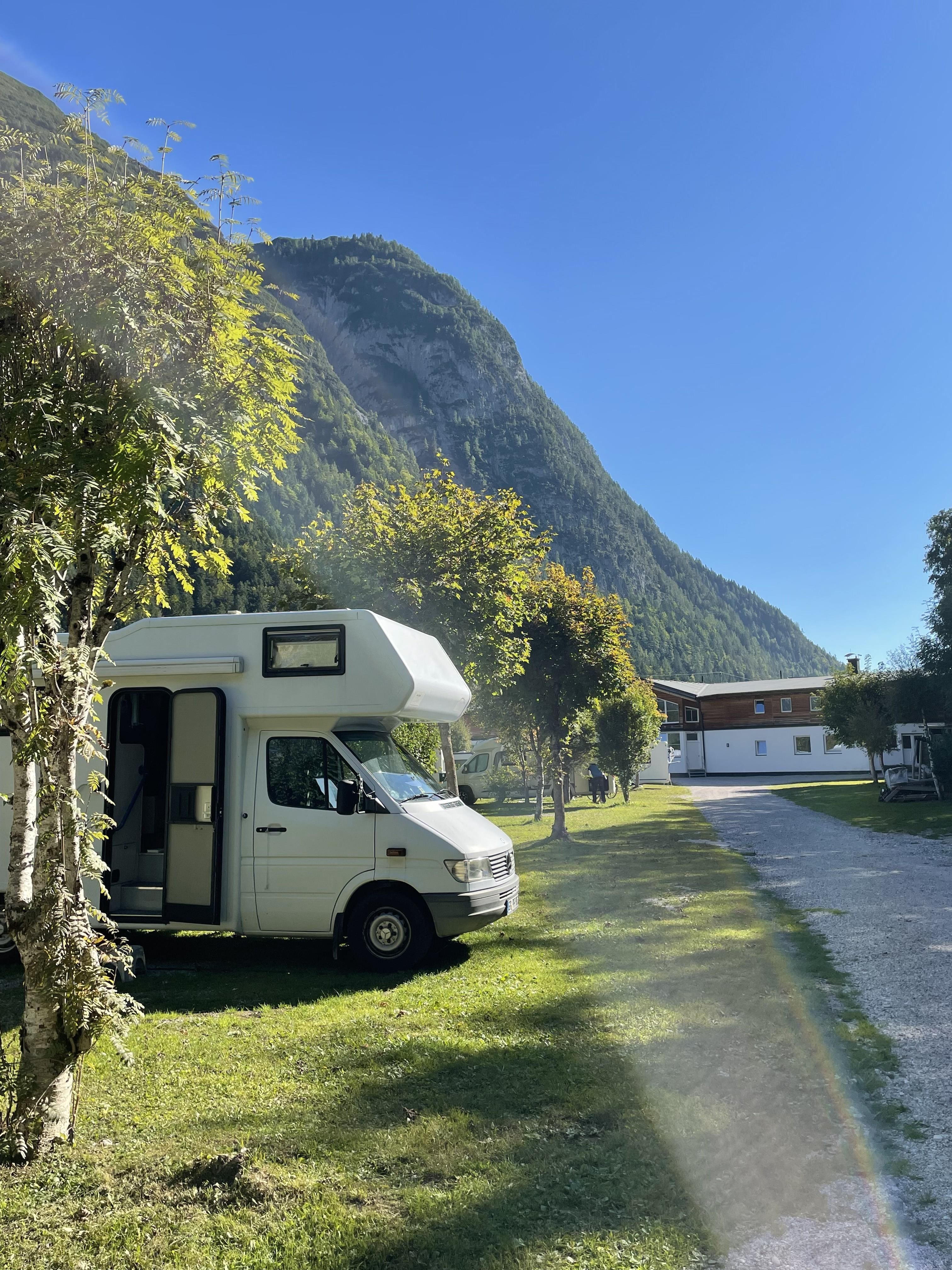A motorhome is parked on a green campsite surrounded by trees. In the background, tall mountains and a clear blue sky can be seen.