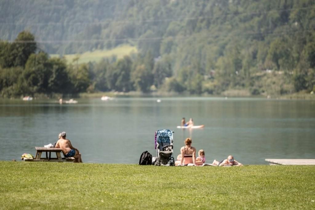 A calm lake with green grass and a few people relaxing. In the background, paddlers can be seen on the water.