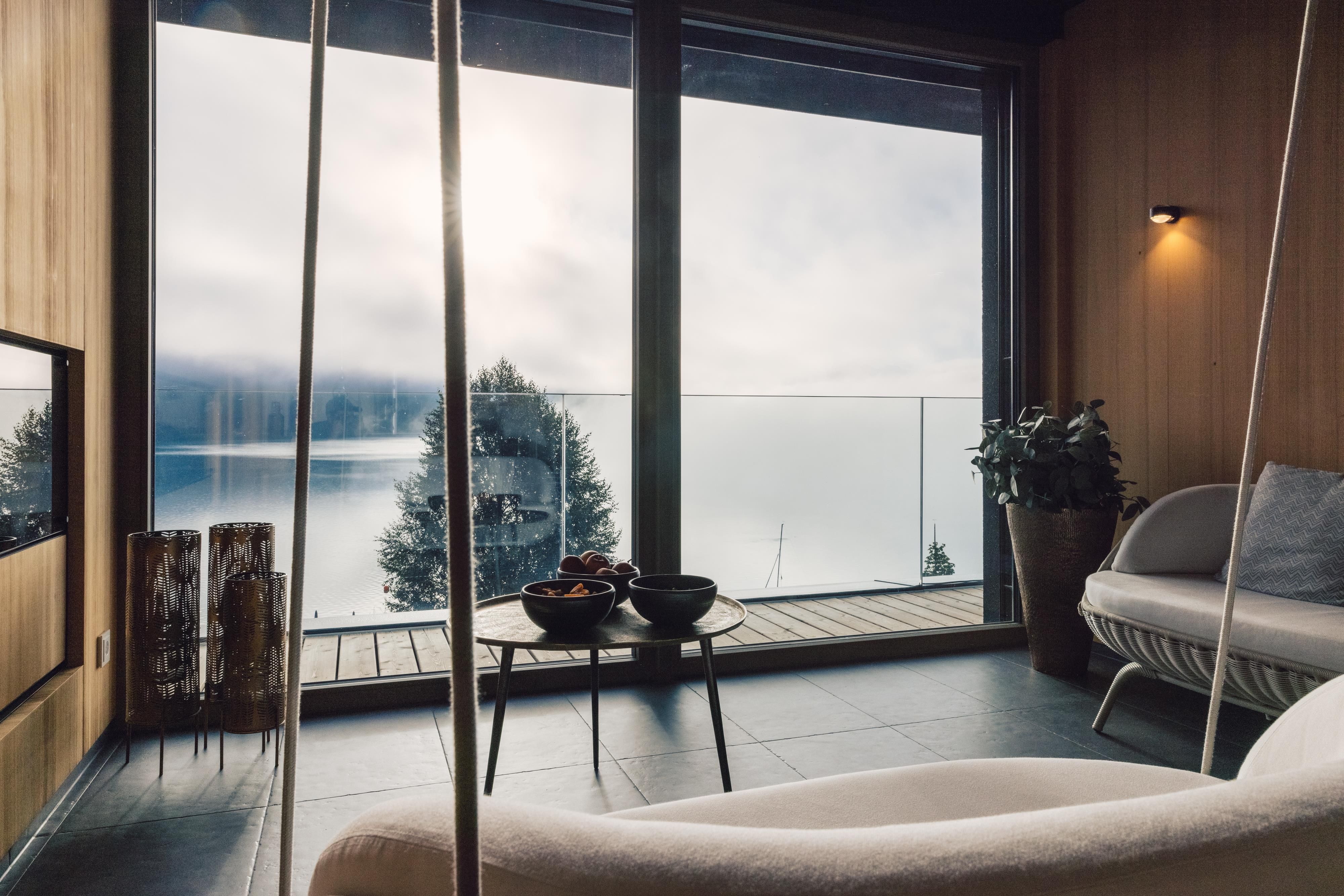 A stylish living room with large windows overlooking a tranquil lake. The wooden walls and modern furniture create a cozy atmosphere.