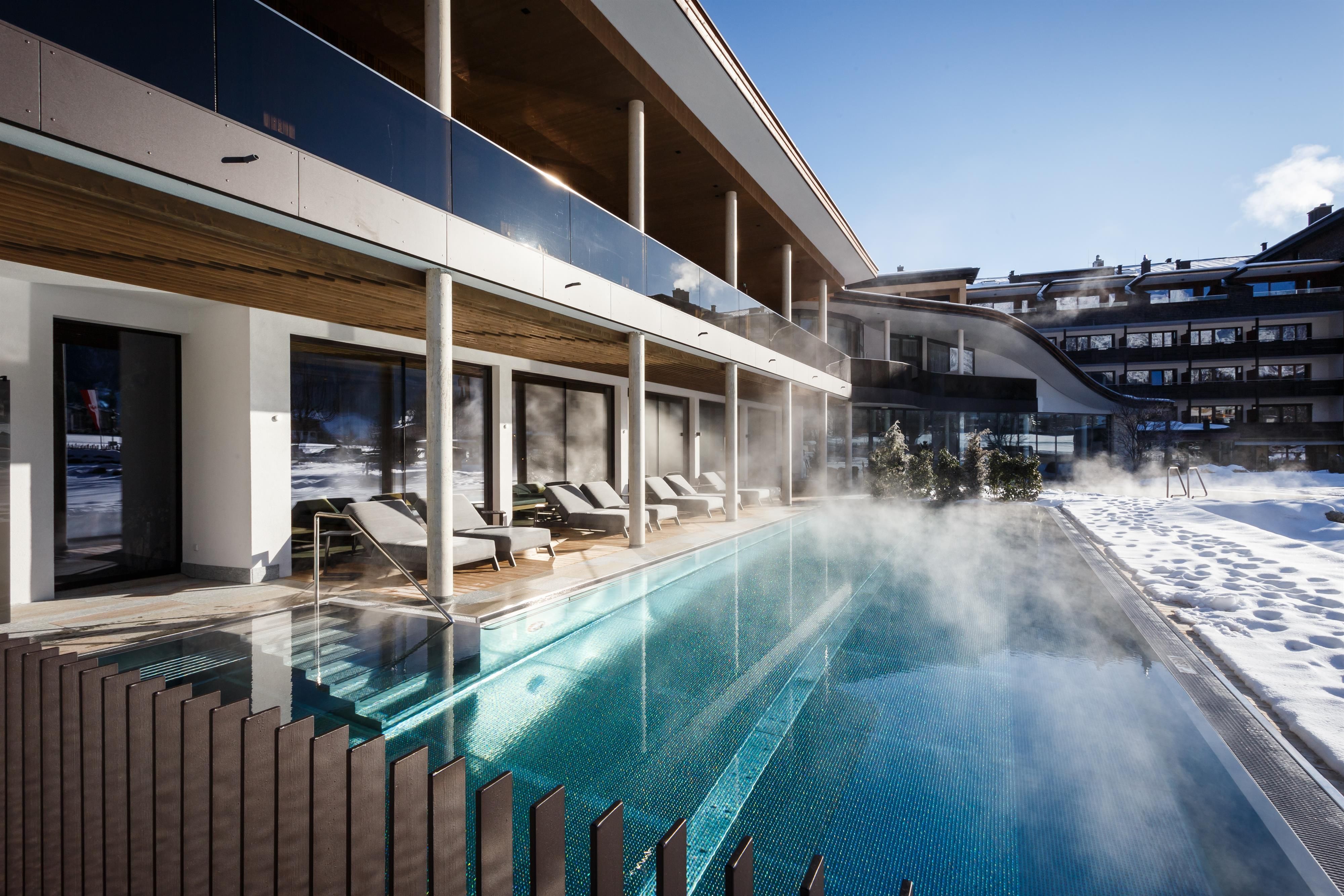 A modern hotel with a heated outdoor pool and comfortable loungers. In the background, snow can be seen, creating a cozy winter atmosphere.