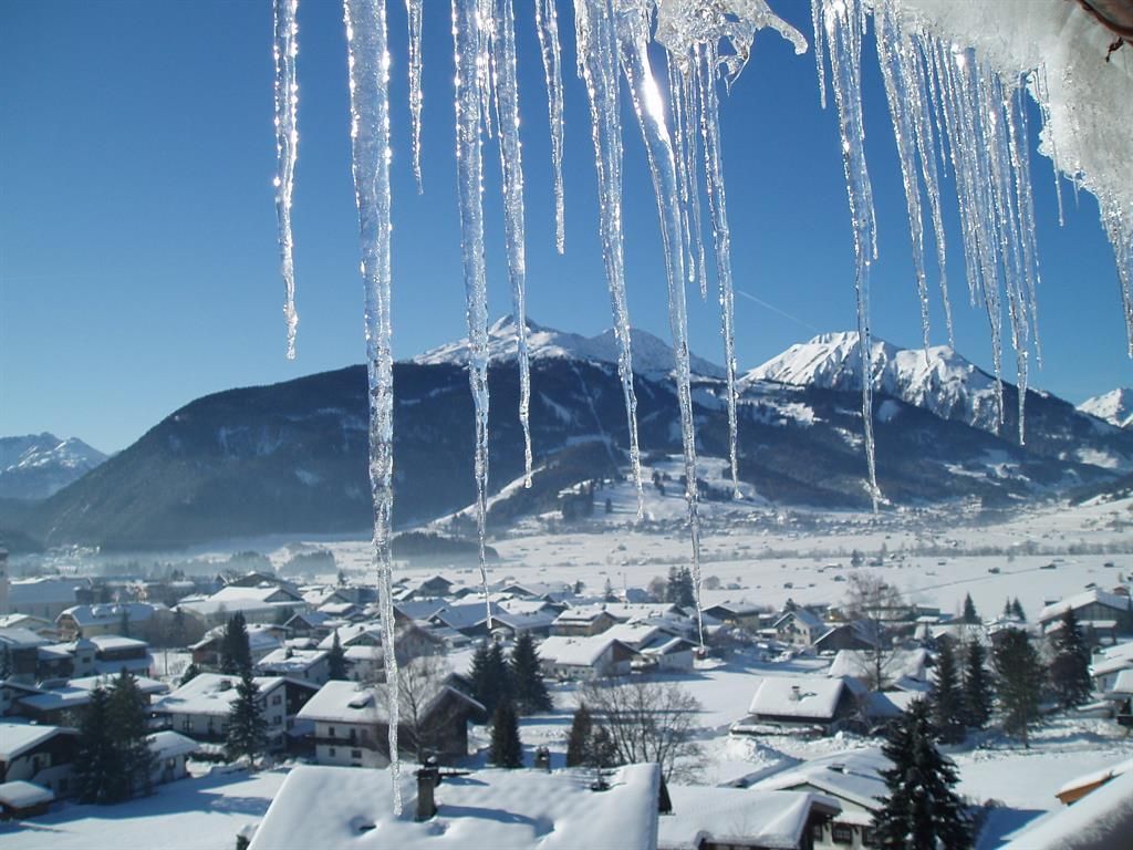 A winter village with snow-covered houses and an impressive mountain landscape. Icicles hang from a roof and sparkle in the sun.