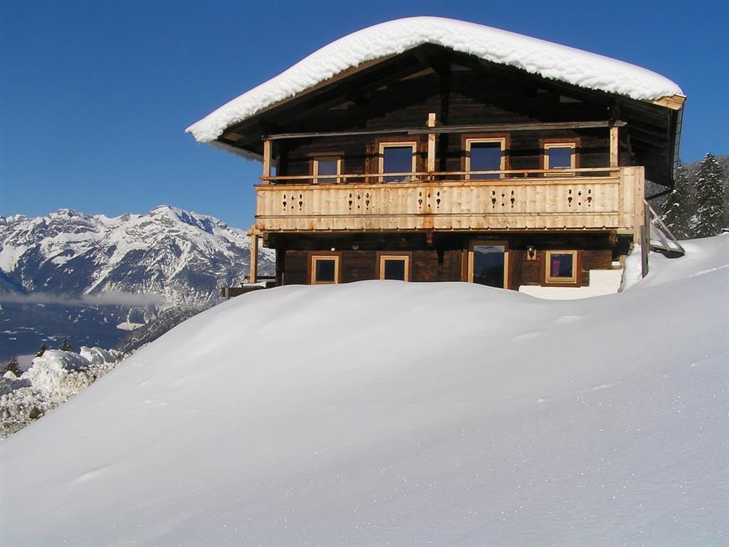 A cozy wooden house in the snow with a large balcony. In the background, majestic mountains are visible under a clear blue sky.