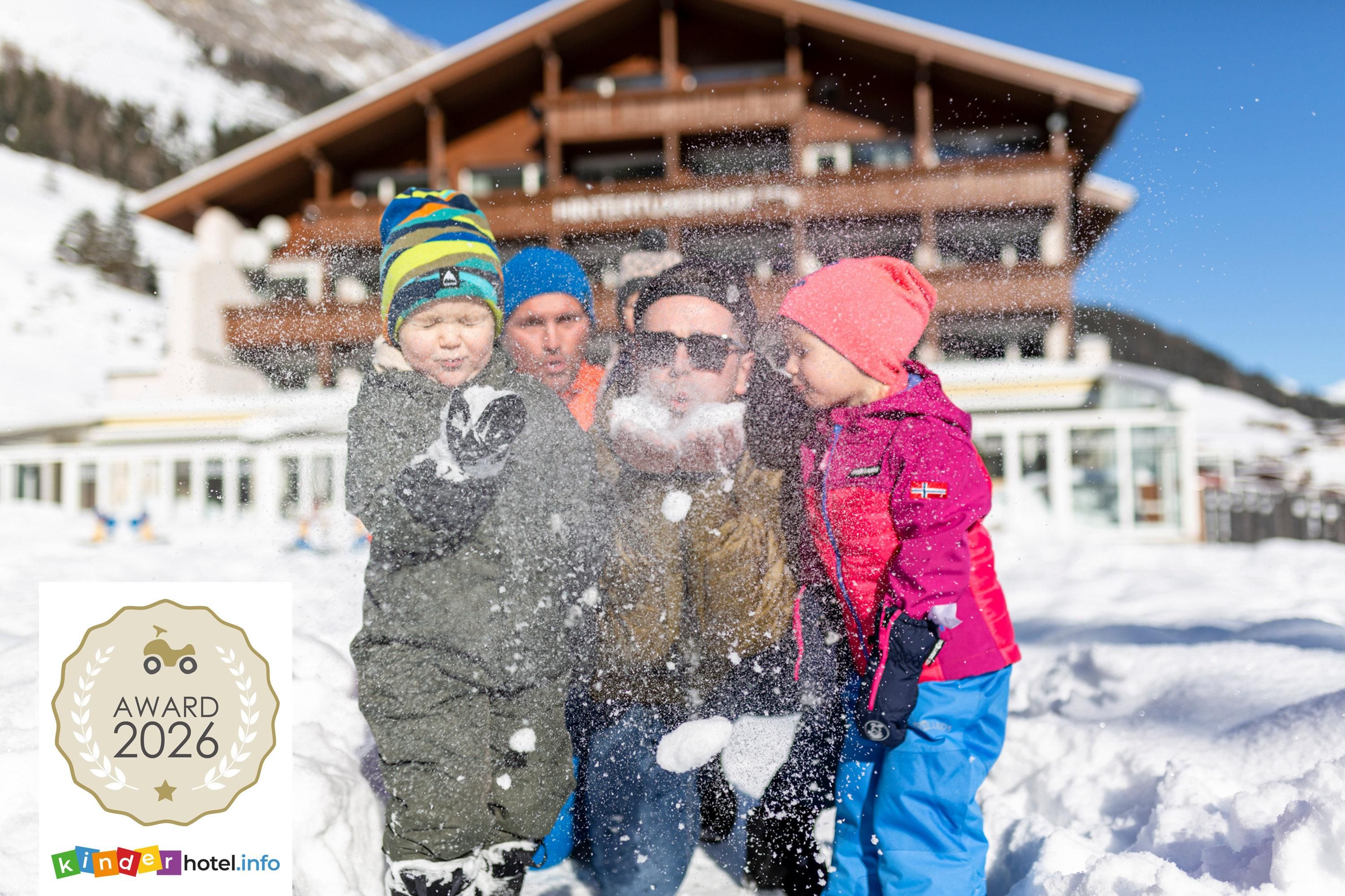 A family plays in the snow in front of a large wooden building. They laugh and throw snow into the air while the sun shines.