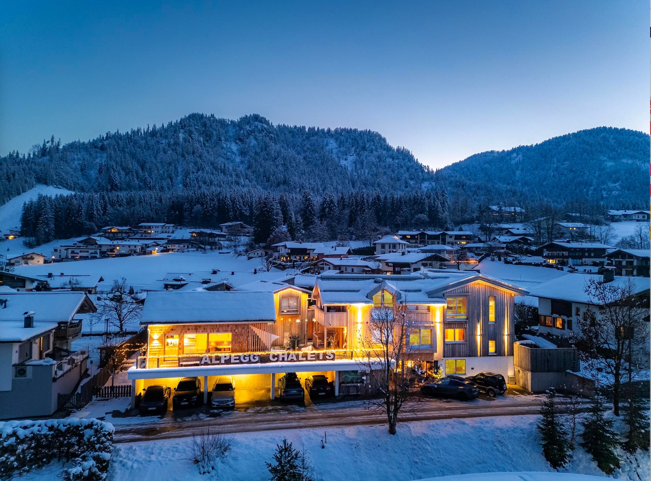 A cozy hotel in a snowy landscape, surrounded by mountains. The lighting of the house radiates warmth into the cold surroundings.