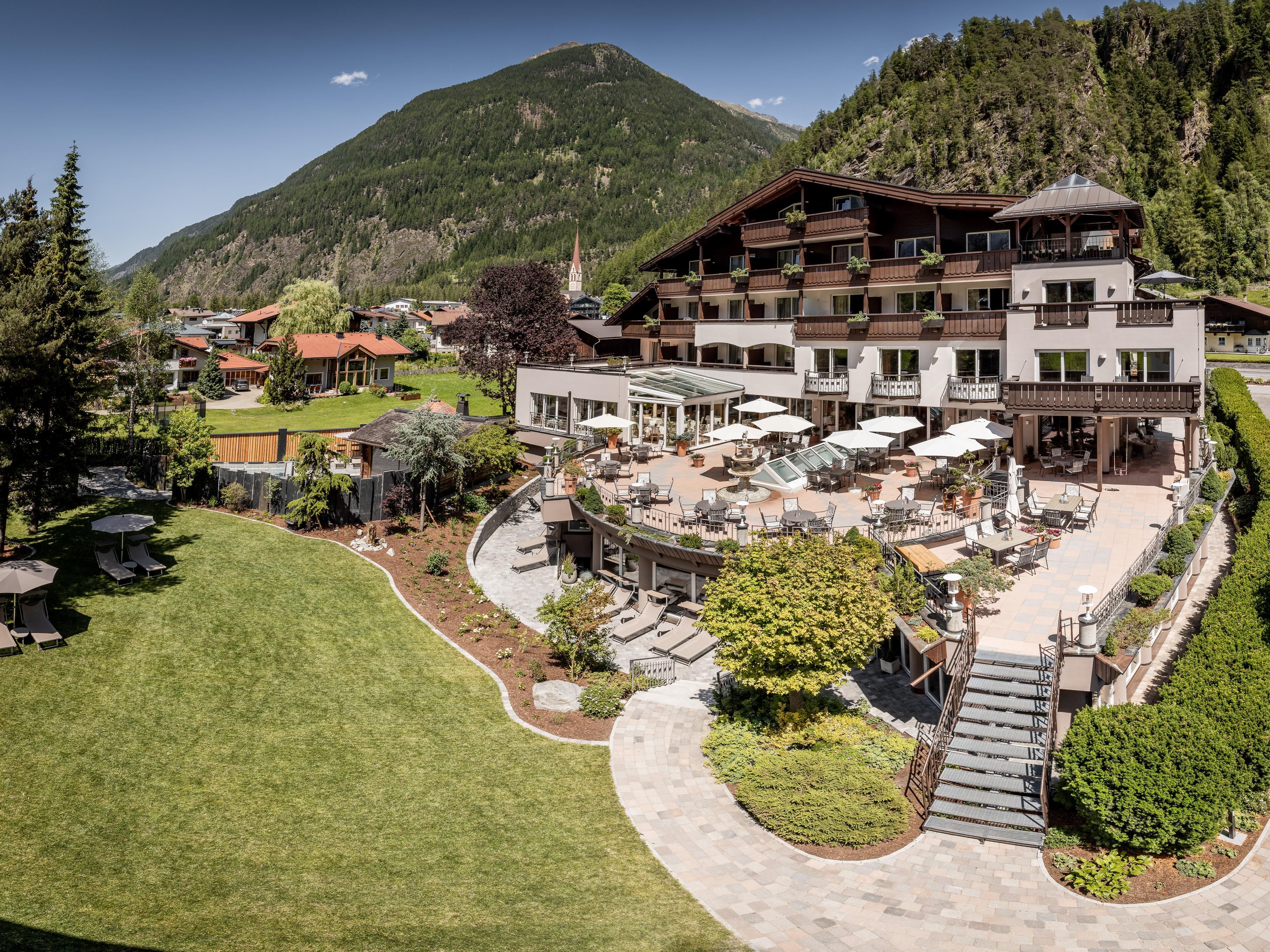 A beautiful hotel with a large garden and a terrace. In the background, green mountains and a clear sky can be seen.