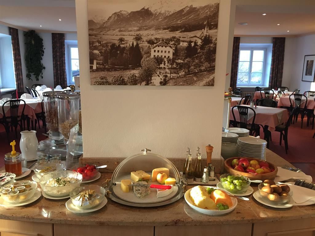 A rich breakfast buffet with various types of cheese, fresh fruit, and vegetables. In the background, there is a large picture of a mountain landscape.