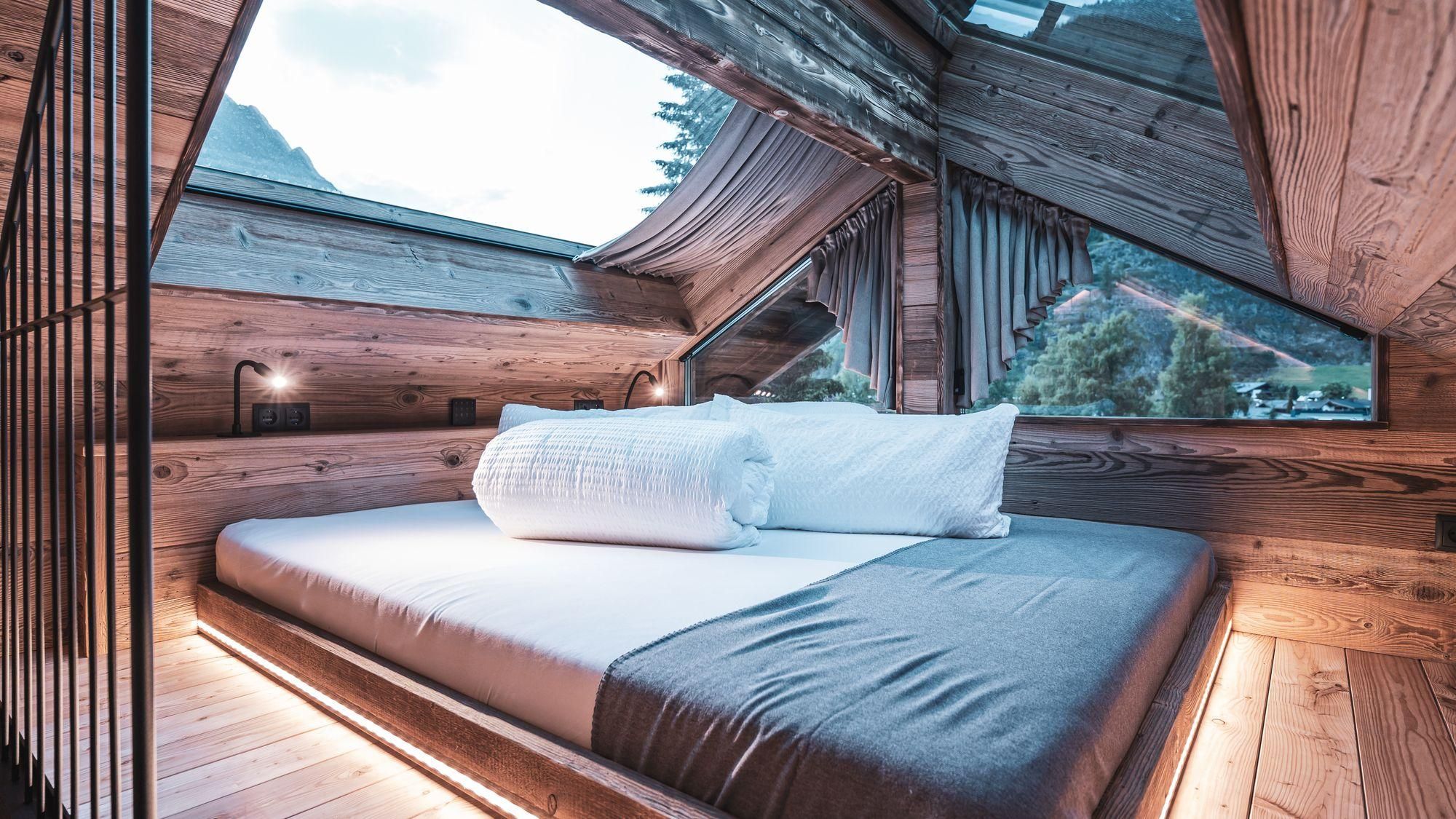 A modern bedroom with a wooden ceiling and large windows. The bed is cozy and inviting, surrounded by natural light.